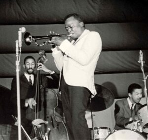 Czarno-białe zdjęcie z 1963 roku: Miles Davis w białej marynarce gra na trąbce na scenie w Antibes; obok Ron Carter w garniturze gra na kontrabasie, a Tony Williams w garniturze siedzi przy perkusji.